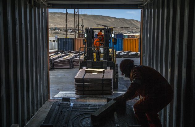 Dentro de contêiner, empilhadeira carrega placas de cobre. Um trabalhador auxilia. Ao fundo, paisagem industrial árida.