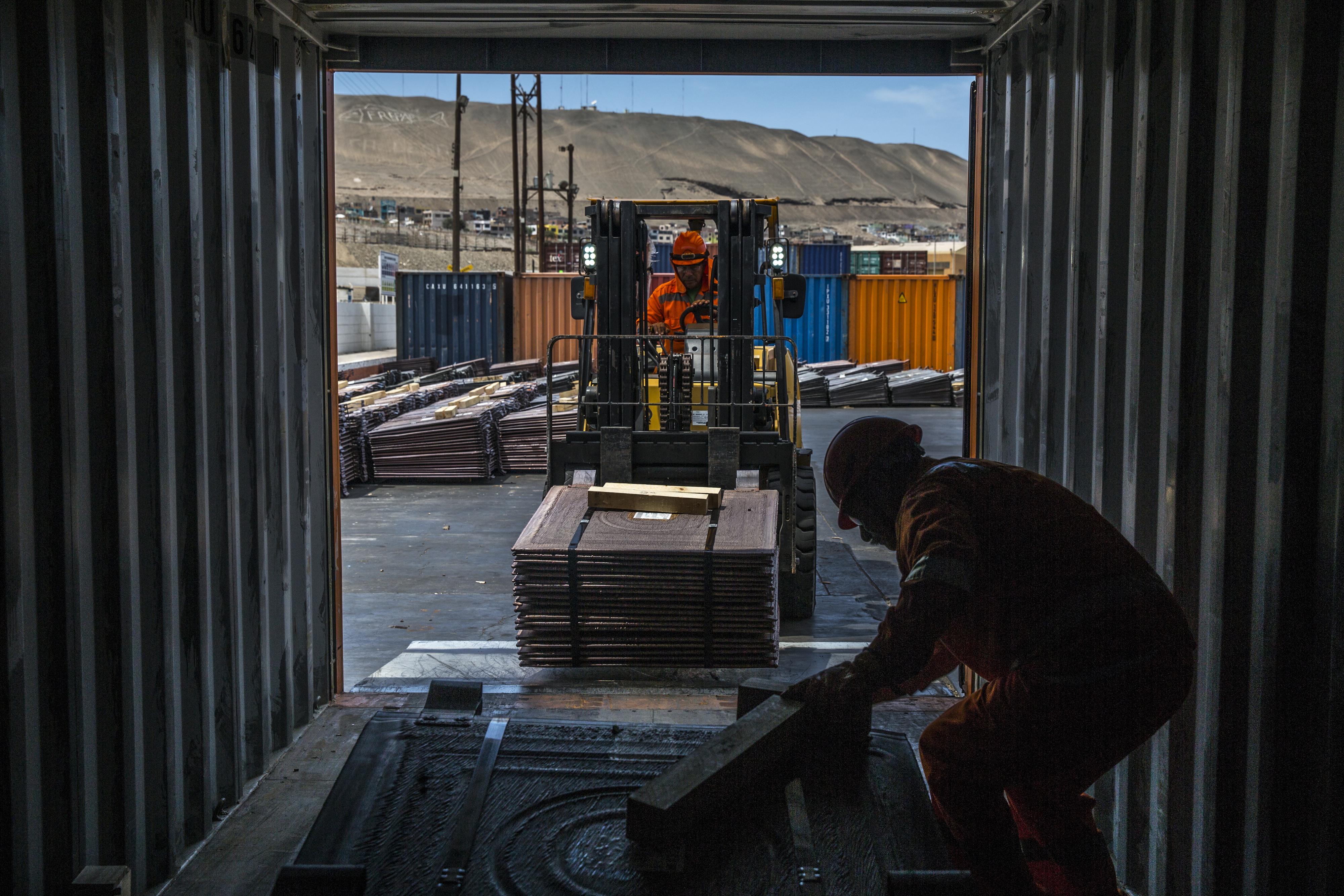 Dentro de contêiner, empilhadeira carrega placas de cobre. Um trabalhador auxilia. Ao fundo, paisagem industrial árida.