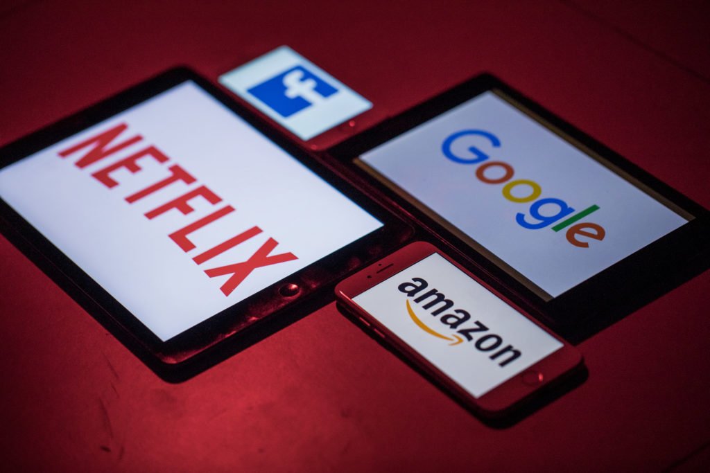 Quatro dispositivos eletrônicos exibindo logotipos de Netflix, Google, Facebook e Amazon sobre fundo vermelho.