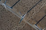 Vista aérea de turbinas eólicas e pás no solo em paisagem árida, com estradas de terra e sombras longas.