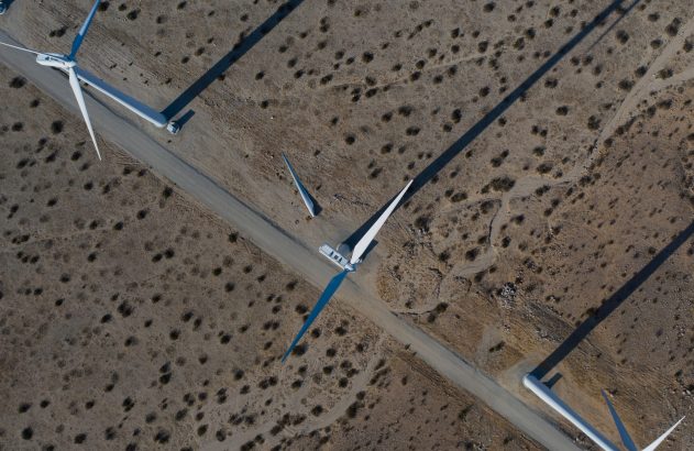 Vista aérea de turbinas eólicas e pás no solo em paisagem árida, com estradas de terra e sombras longas.