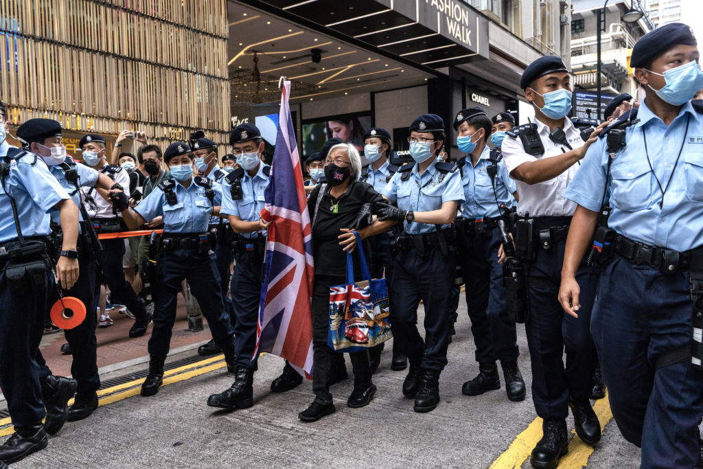 Homem ergue bandeira britânica, cercado por policiais mascarados em rua.