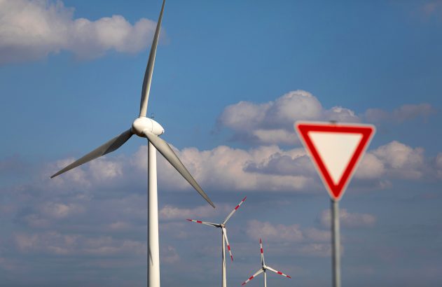 Turbina eólica com pás girando e placa de “Dê a Preferência” sob céu azul. Outras turbinas ao fundo.