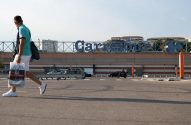 Homem com sacola atravessa estacionamento. Ao fundo, loja Carrefour com letreiro azul e veículos.