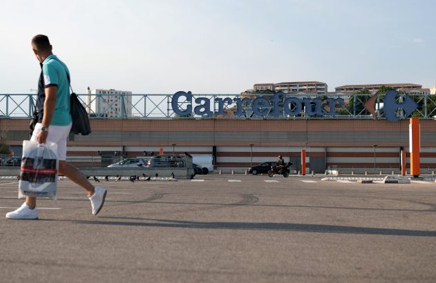 Homem com sacola atravessa estacionamento. Ao fundo, loja Carrefour com letreiro azul e veículos.