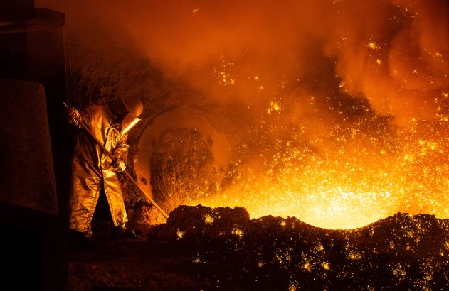 Trabalhador em equipamento protetor lida com metal fundido e faíscas intensas em siderúrgica.