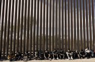 Homens sentados no chão diante de um muro de fronteira alto de metal com barras verticais.