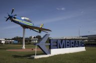 Avião de exposição nas cores do Brasil sobre mastro, ao lado do letreiro "EMBRAER".