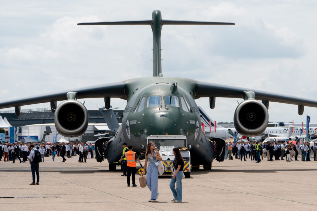 Avião de transporte militar Embraer C-390 em exposição, visto de frente, com público e outras aeronaves no fundo.