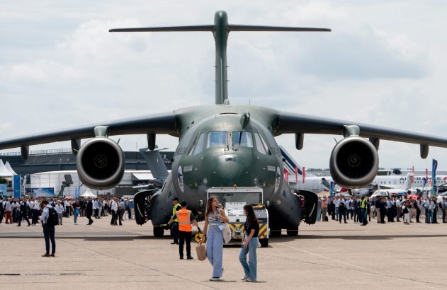 Avião de transporte militar Embraer C-390 em exposição, visto de frente, com público e outras aeronaves no fundo.
