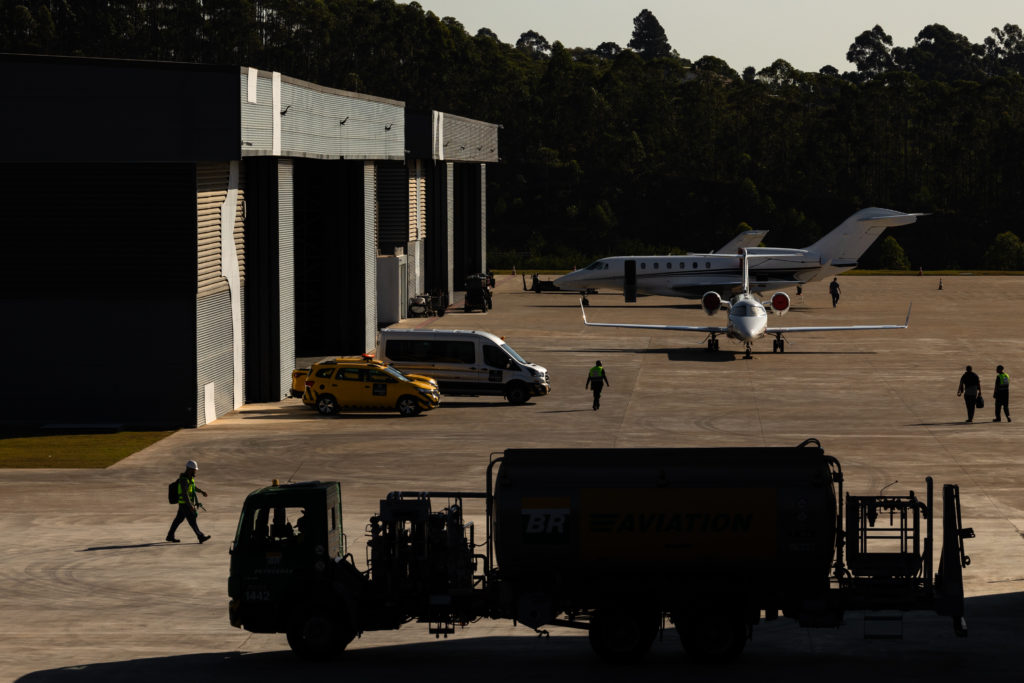 Pátio de aeroporto com jatos executivos, hangares e veículos de apoio. Funcionários caminham sob forte luz solar.