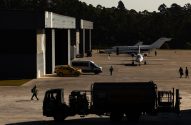 Pátio de aeroporto com jatos executivos, hangares e veículos de apoio. Funcionários caminham sob forte luz solar.