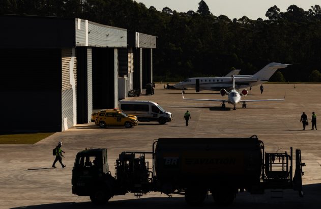 Pátio de aeroporto com jatos executivos, hangares e veículos de apoio. Funcionários caminham sob forte luz solar.