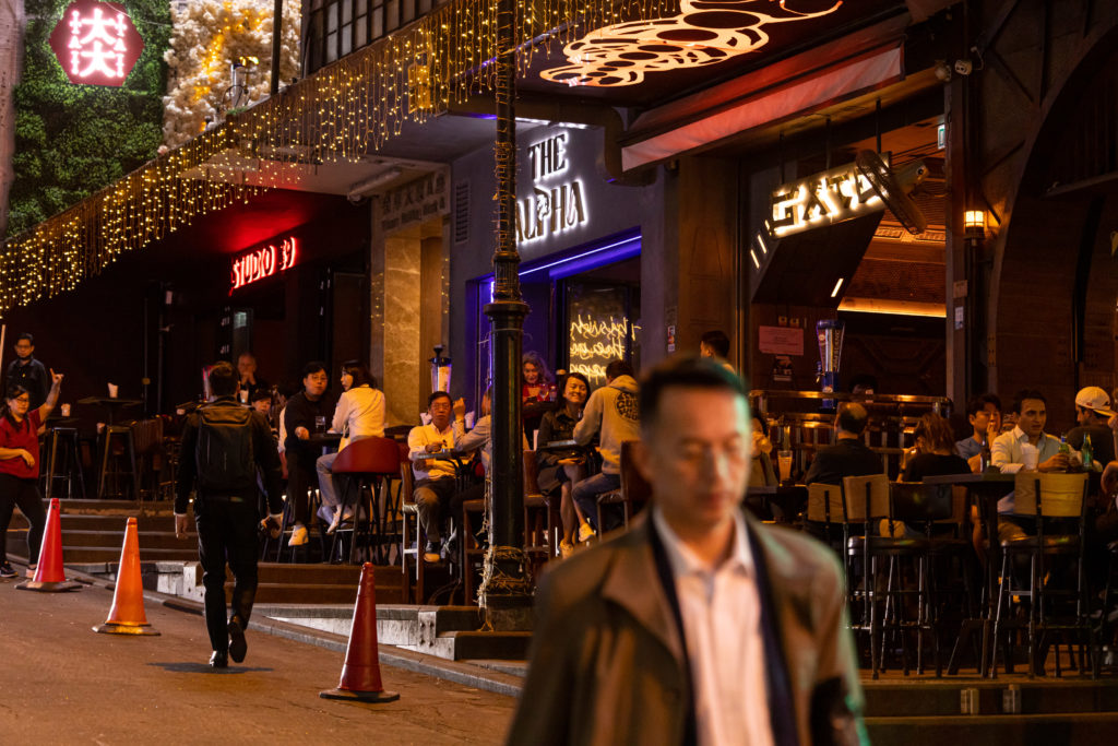 Rua noturna movimentada com bares e luzes neon, pessoas socializando e um homem caminhando.