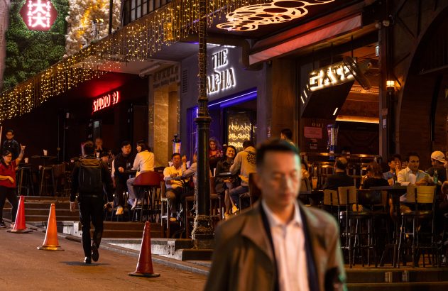 Rua noturna movimentada com bares e luzes neon, pessoas socializando e um homem caminhando.