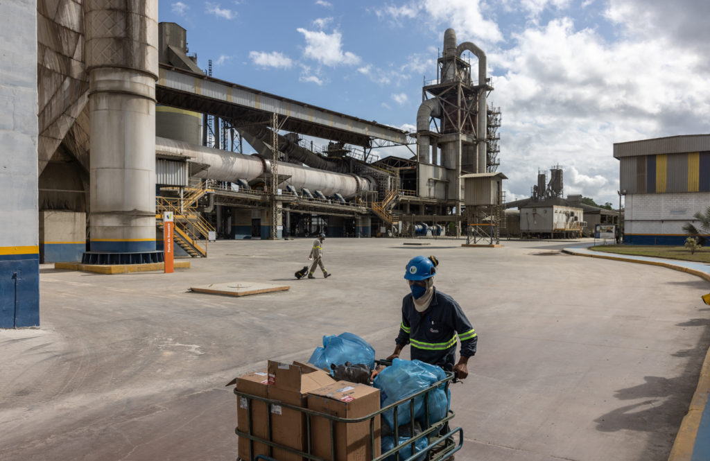 Homem empurra carrinho com carga em pátio de fábrica industrial; outro trabalhador anda ao fundo.