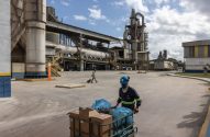 Homem empurra carrinho com carga em pátio de fábrica industrial; outro trabalhador anda ao fundo.
