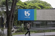 Homem caminha em frente a prédio da Sabesp, com logo azul e verde. Árvores ao redor.