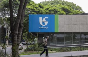 Homem caminha em frente a prédio da Sabesp, com logo azul e verde. Árvores ao redor.