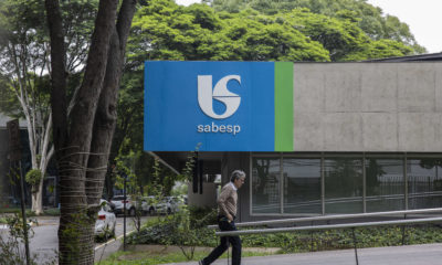 Homem caminha em frente a prédio da Sabesp, com logo azul e verde. Árvores ao redor.