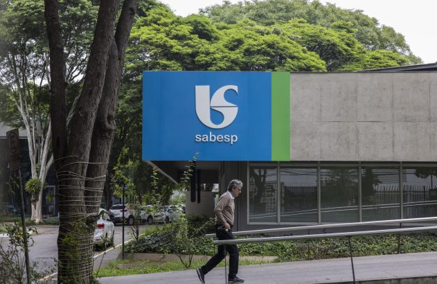 Homem caminha em frente a prédio da Sabesp, com logo azul e verde. Árvores ao redor.