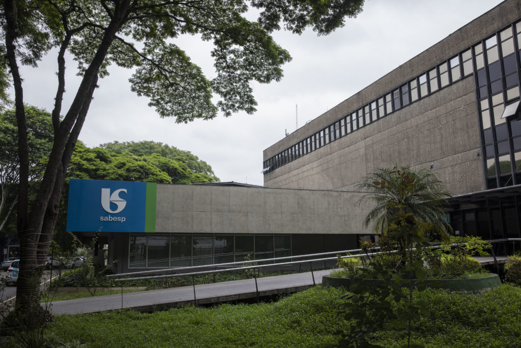 Prédio da Sabesp de concreto, com logotipo azul e verde, janelas, árvores, gramado e uma rampa de acesso sob céu nublado.