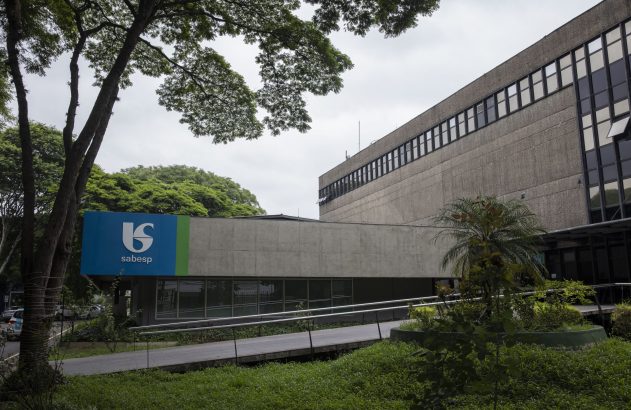 Prédio da Sabesp de concreto, com logotipo azul e verde, janelas, árvores, gramado e uma rampa de acesso sob céu nublado.
