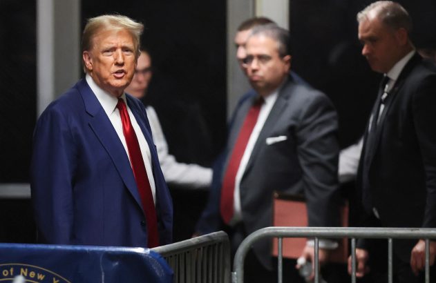 Donald Trump em terno azul e gravata vermelha, caminhando e olhando diretamente para a câmera, com seguranças atrás.