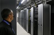 Homem observa data center com fileiras de racks de servidores. Alguns estão abertos, revelando cabos e equipamentos.