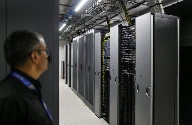 Homem observa data center com fileiras de racks de servidores. Alguns estão abertos, revelando cabos e equipamentos.