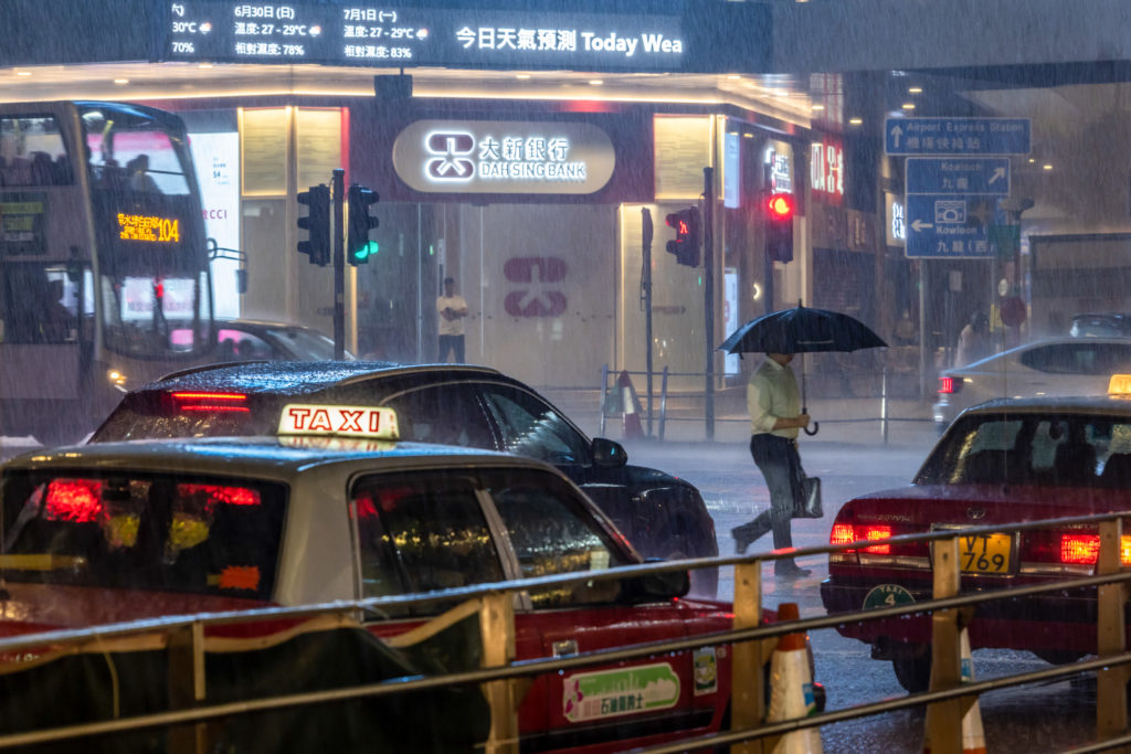 Rua urbana chuvosa, com táxis e um homem de guarda-chuva atravessando. Prédio do banco e painel de tempo ao fundo.