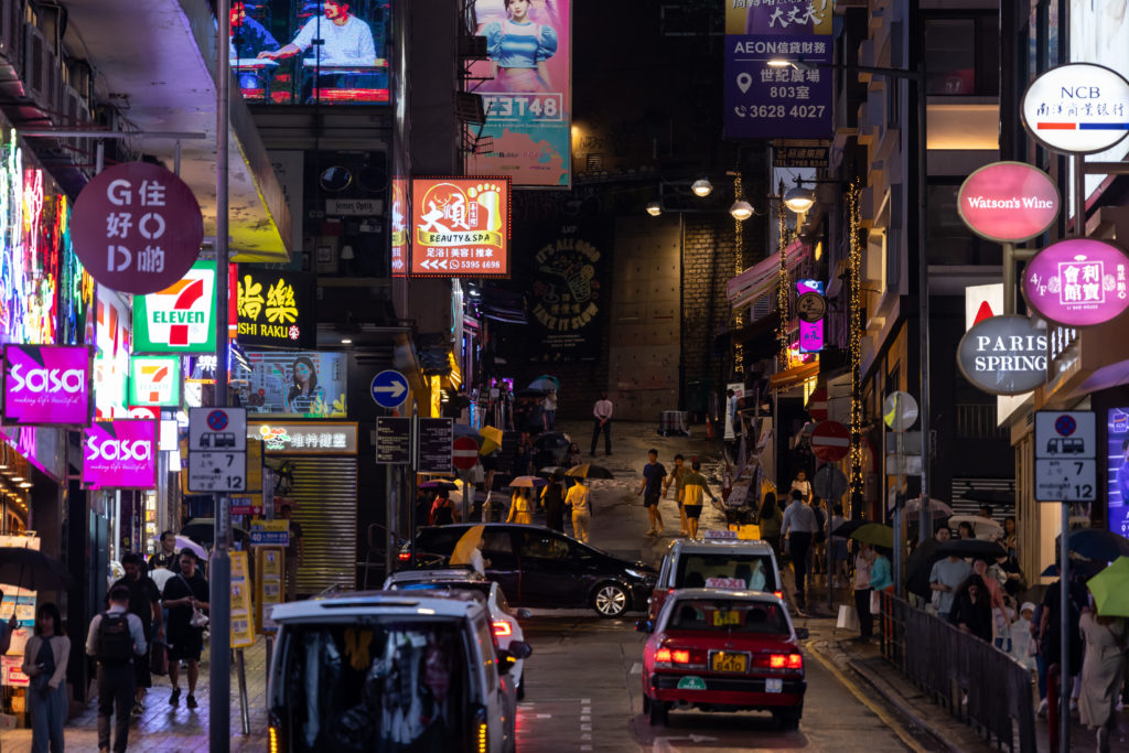 Noite em rua de Hong Kong com letreiros neon vibrantes. Carros e pedestres se movem em meio à iluminação colorida.