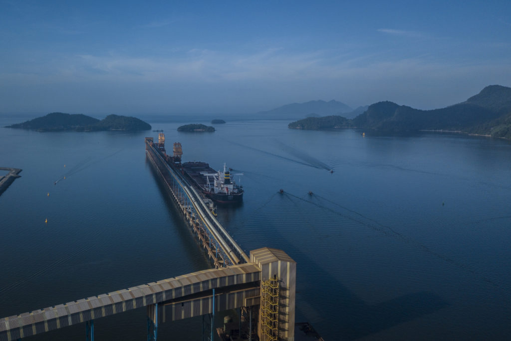 Vista aérea: navio graneleiro em píer de carregamento em baía, com esteiras transportadoras, ilhas e montanhas.