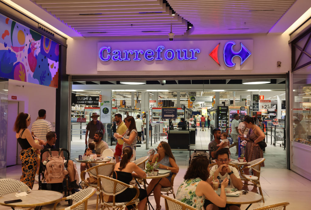 Entrada do Carrefour. Pessoas sentadas em mesas de café e outras entrando e saindo do supermercado.