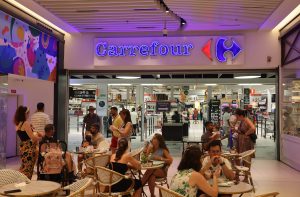Entrada do Carrefour. Pessoas sentadas em mesas de café e outras entrando e saindo do supermercado.