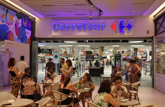 Entrada do Carrefour. Pessoas sentadas em mesas de café e outras entrando e saindo do supermercado.