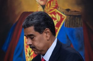 Nicolás Maduro de terno escuro e gravata vermelha caminha, com pintura de Simón Bolívar ao fundo.