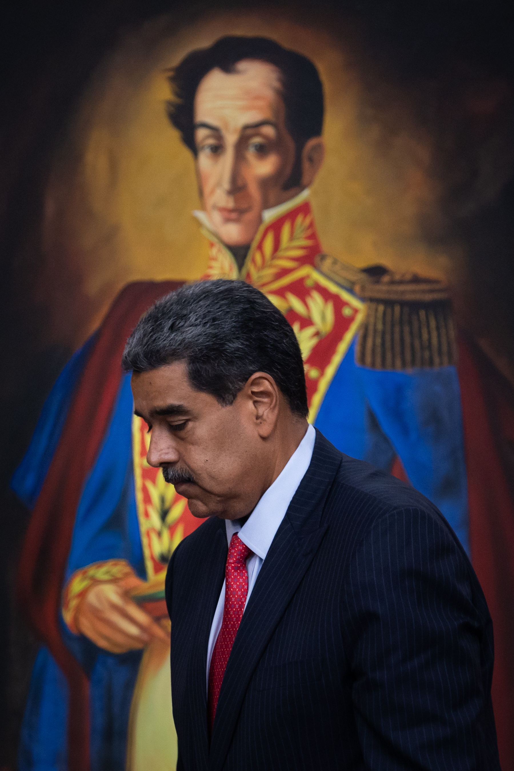 Nicolás Maduro de terno escuro e gravata vermelha caminha, com pintura de Simón Bolívar ao fundo.