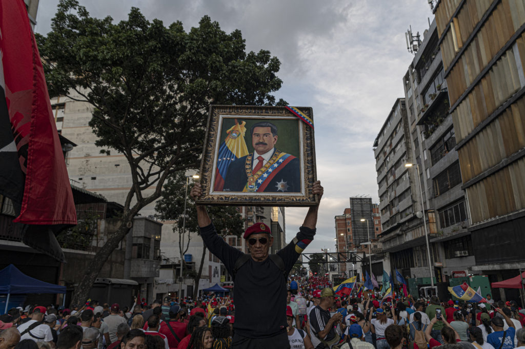 Homem de boina vermelha ergue retrato de Nicolás Maduro em multidão, com bandeiras venezuelanas visíveis.