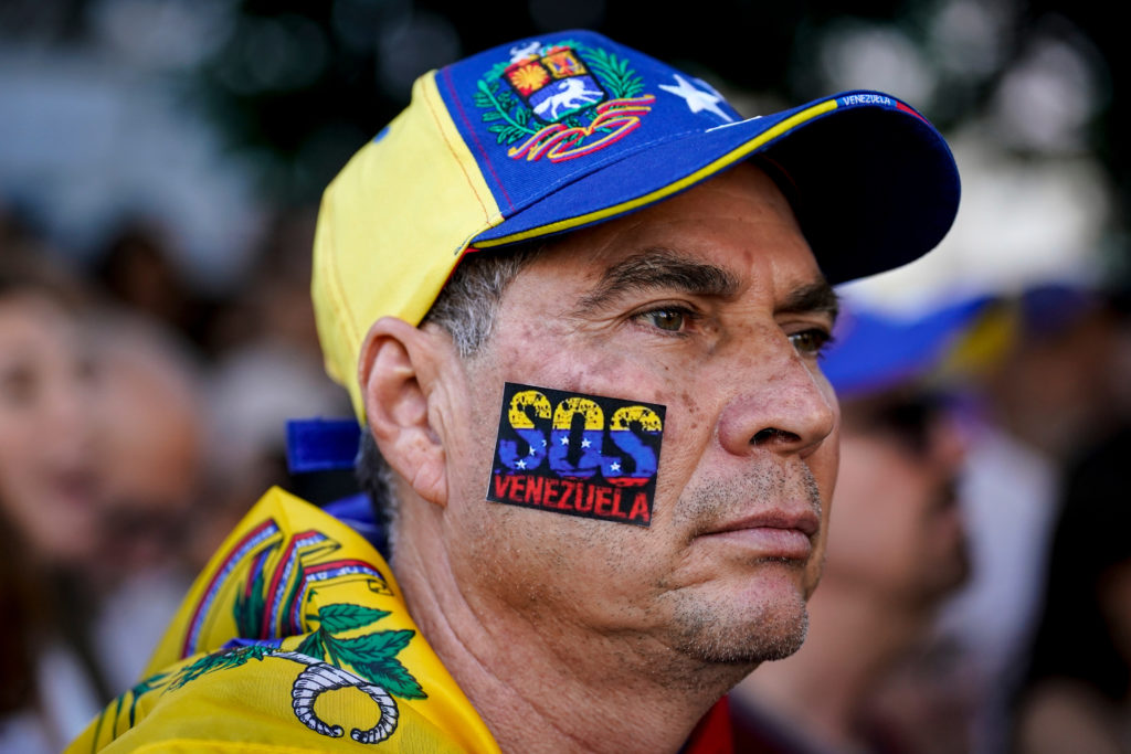 Homem de boné venezuelano e camisa, com adesivo "SOS Venezuela" no rosto, olhando para cima com expressão séria.