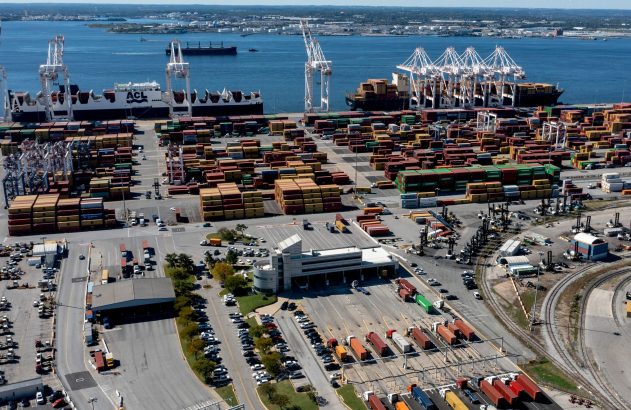 Vista aérea de porto de contêineres com navios, guindastes, pilhas de contêineres coloridos e caminhões.