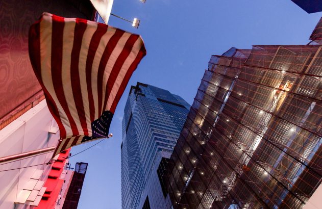 Bandeira dos EUA pendurada. Prédios altos, um coberto por andaimes e rede, elevam-se sob o céu azul ao entardecer na cidade.