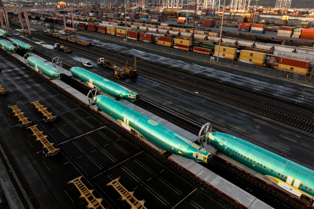 Fuselagens de avião verde-azuladas sendo transportadas por trem em um pátio ferroviário movimentado.