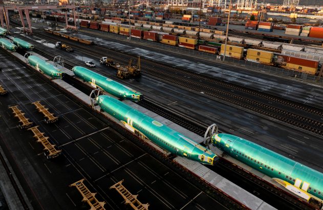 Fuselagens de avião verde-azuladas sendo transportadas por trem em um pátio ferroviário movimentado.