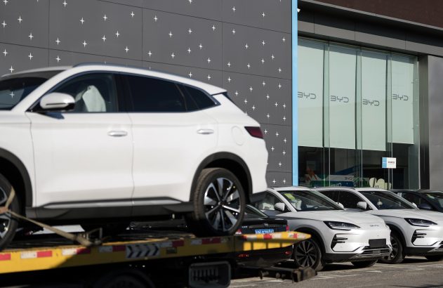 Carros SUV brancos, um sendo transportado em caminhão-guincho, outros estacionados em frente a uma concessionária BYD.