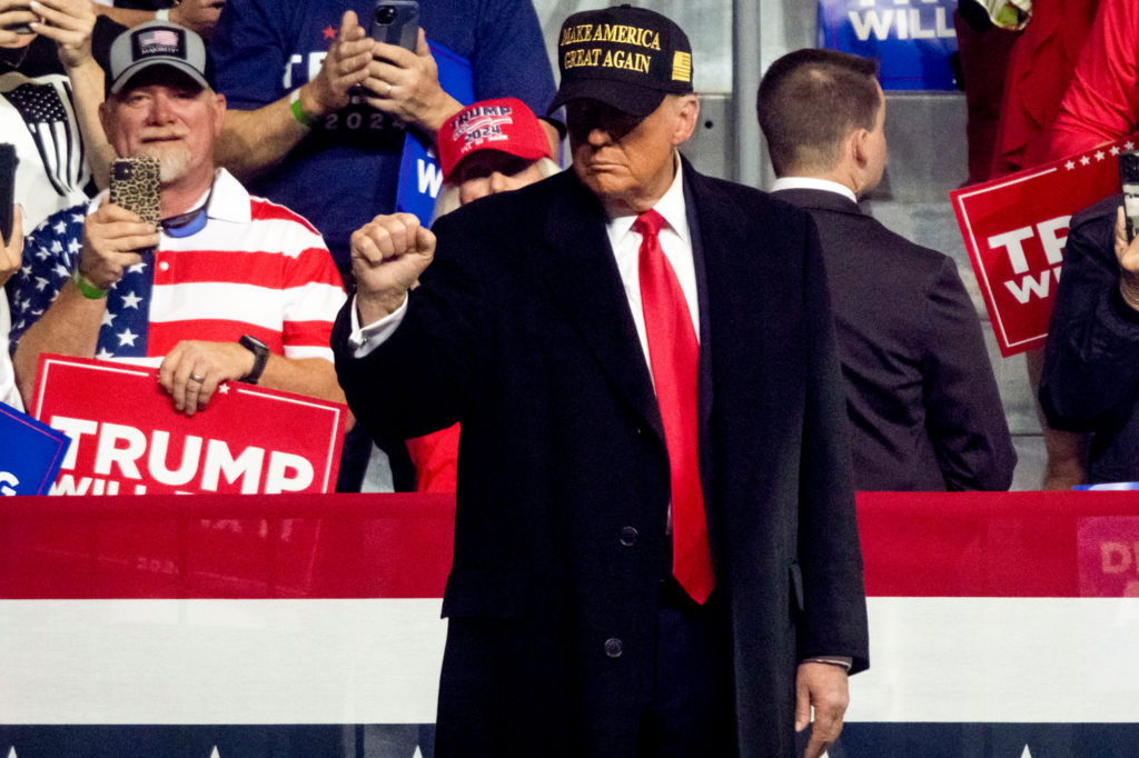 Donald Trump, de chapéu MAGA, punho erguido, em palco de evento político. Apoiadores com placas e camisas ao fundo.