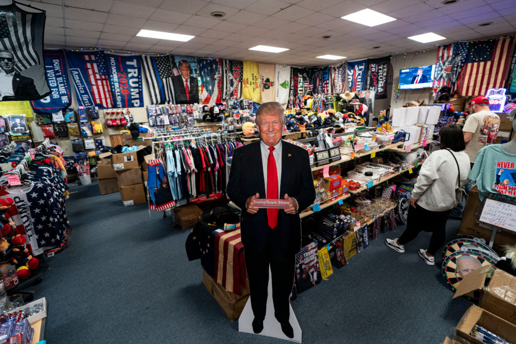 Loja temática de Donald Trump com bandeiras, roupas e um recorte de papelão do ex-presidente. Clientes compram.