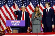 Donald Trump, Melania e Barron em palco com bandeiras dos EUA. Trump acena, Melania aplaude. Pódio: 'Trump Will Fix It 2024'.