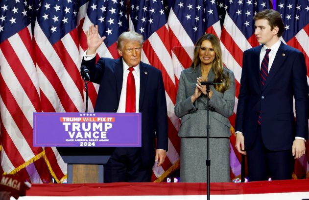Donald Trump, Melania e Barron em palco com bandeiras dos EUA. Trump acena, Melania aplaude. Pódio: 'Trump Will Fix It 2024'.