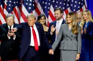 Donald Trump aponta e segura a mão de Melania Trump. Ivanka e outros aplaudem em evento com bandeiras dos EUA.